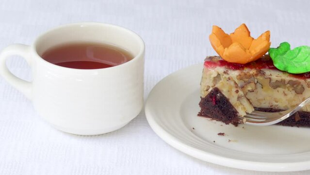 pastry piece slice of cake rectangle shape on white plate and black tea in cup.strawberry cake walnuts orange flower green leaf or pink macaron decoration.fork cutting the desert.tasty morning delight