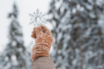hand in mitten holding decorative transparent snowflake, winter forest natural background.