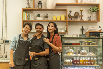 Three young ladies express their confidence in running a startup coffee shop. Three ladies stand in front of a coffee counter ready to serve their customers. Three baristas show positive body gesture.