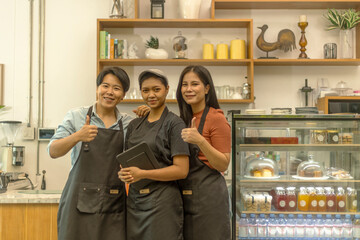Three young ladies express their confidence in running a startup coffee shop. Three ladies stand in front of a coffee counter ready to serve their customers. Three baristas show positive body gesture.