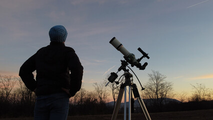 Silhouette of a man, telescope and countryside under evening skies.