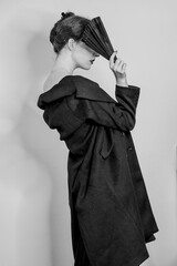 Obraz premium Vertical grayscale shot of an elegant female posing in a slip dress, trench coat, and a fan
