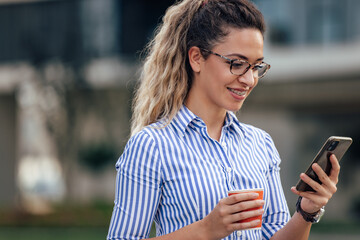 Smiling girl chatting with someone over the phone, holding a cup of coffee, walking outdoor.