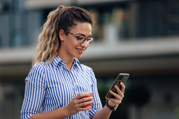 Side view of a smiling girl, using a phone and holding cup of coffee.