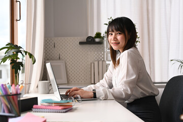 Pretty young woman employee in white shirt sitting front of laptop computer and smiling to camera
