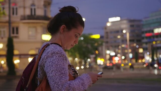 Woman tourist checking her mobile phone at night in central city square. Female standing in middle of plaza with city lights and cars in background during blue hour or dusk holding smartphone