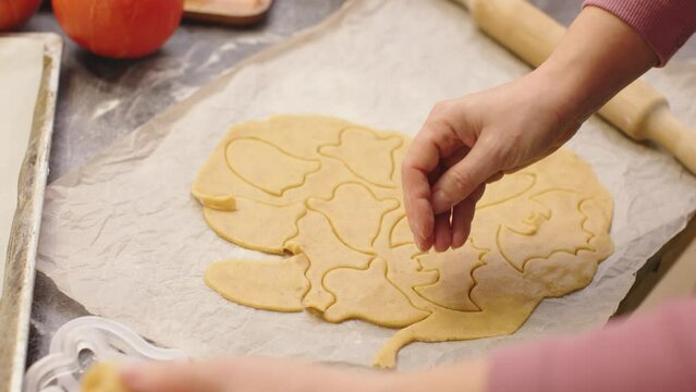 hands cutting out raw halloween sweet cookies ghost shape form from dough pastry put homemade biscuits on baking sheet bakery pan tray. celebrating October happy halloween holiday party trick or treat
