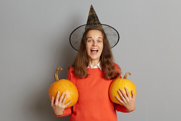 Indoor shot of excited amazed Caucasian woman wearing sweater and witch hat standing with orange...