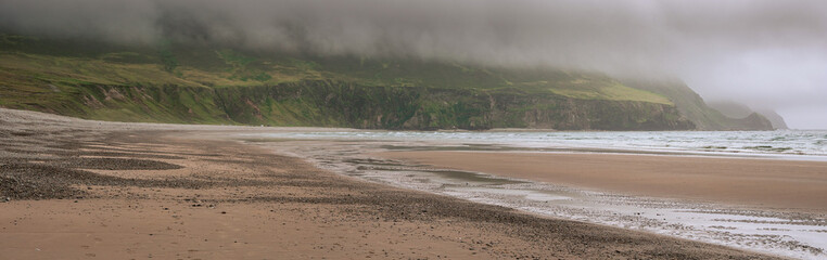 Panorama image of stunning nature scenery and Keem beach, county Mayo, Ireland. Popular travel area with surfing and sightseeing. Irish landscape. Low clouds, rainy day. Stormy weather.