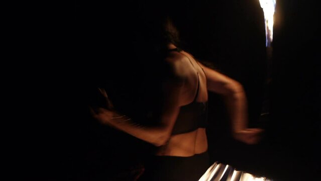 Asian woman spinning fire ropes in dark, filmed as tight medium shot from backside