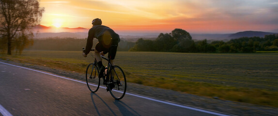  Cyclist riding his bicycle on an empty rural road at dawn, epic sunrise in the background
