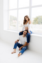 Young happy pregnant couple in love barefoot sitting on windowsill. Laughing and smiling man and woman. Happy pregnancy concept