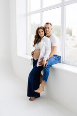 Happy family is having fun while sitting on the windowsill by the panoramic window. Young woman is pregnant, man hugs beloved. Little girl fooling around parents, dancing.