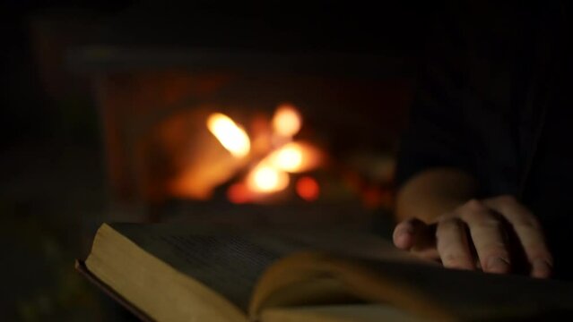 A man reading and turning the pages of a book close to an open fire in a fireplace in a dark room