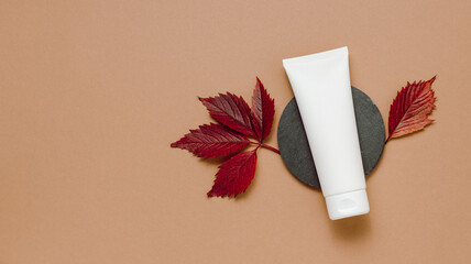 White tube of cream with autumn leaves top view, brown background, mockup cream tube, seasonal skincare, facial cream or moisturizing lotion, minimal aesthetic concept, banner size