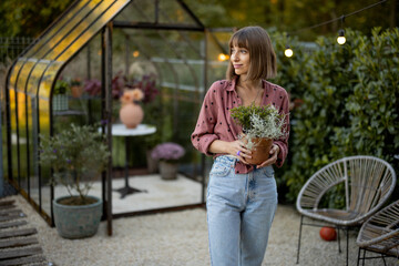 Young woman gardener carries flower pot with herbs on backyard with a beautiful glasshouse for plants on background