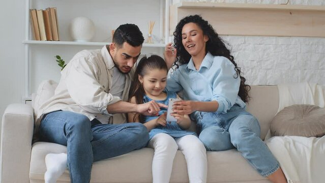 Hispanic Indian multiethnic multiracial family on couch parent mother and father teach kid using phone take photos video with funny masks. Mom and dad with daughter fun laughing child showing tongue