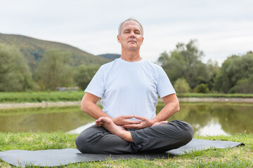 Naklejka na ściany i meble An elderly fit man is sitting in meditation in the lotus position on a yoga mat in a forest clearing in the open air