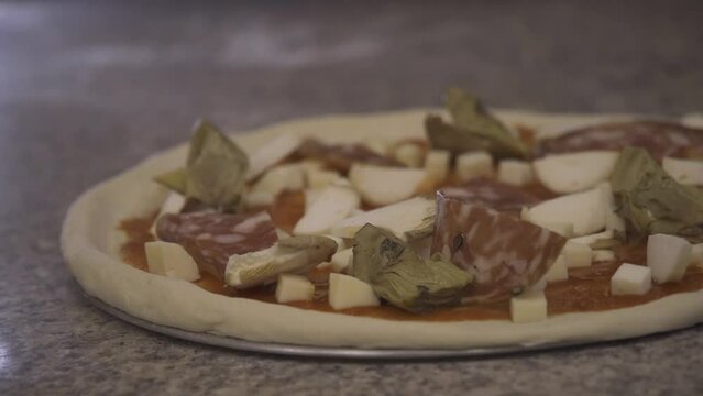 Pizzaiolo prepares Capricciosa pizza on a marble countertop at a restaurant. A professional chef adds artichokes and olives on the yeast pizza dough covered with tomato sauce, cheese, salami and ham.