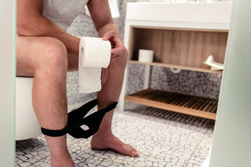 A man holding a roll of toilet tissue in a bathroom, sitting on toilet at morning. Hygiene at...