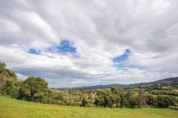 Fototapeta na wymiar Cloudscape over the hills in the Montefeltro. The village of Case Bernardi near Pesaro and Urbino in the Marche region of Italy appears through the green trees