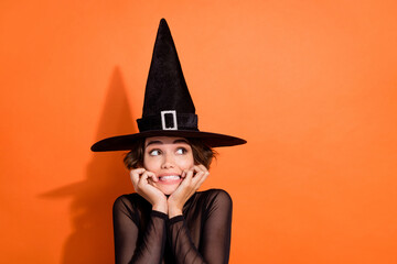 Photo of charming young lady look empty space frighened bite nails wear stylish black halloween witch clothes isolated on orange background