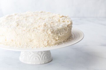 A coconut covered cake on a white pedestal stand against a white background.