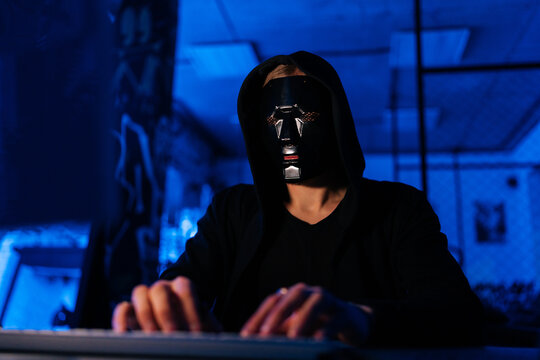 Dangerous unrecognizable hacker man wearing in black mask and sweatshirt hacking online web site or engaging hacking into security systems, using laptop and desktop sitting at desk in dark room.