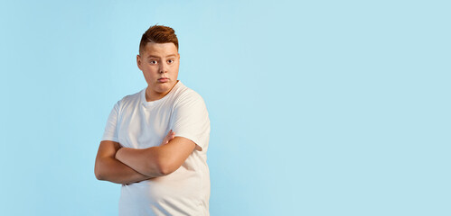 Portrait of teen boy, overweight model in white shirt standing with crossed arms isolated over blue...
