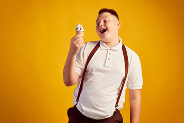 Half-length portrait of happy overweight boy in white t-shirt tasting sweet ice-cream isolated on orange color background. Fast food, taste, body positive, emotions