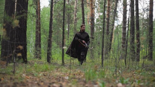 Medieval scout with large bow looks for enemies footprints in forest slow motion. Old soldier in chain mail walks across woodland. Historical reenacting