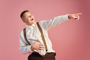 Excited big obese teen boy in white shirt and shorts with suspenders laughing isolated over pink...