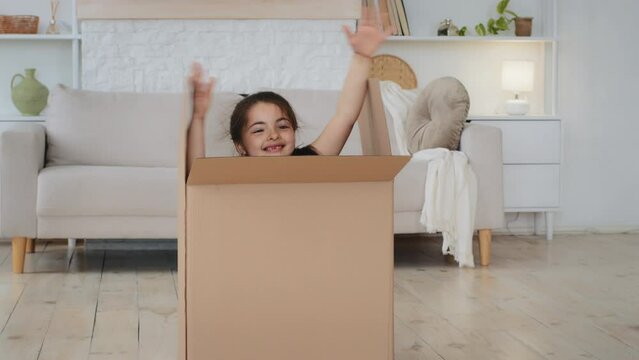 Caucasian child girl daughter make surprise in living room in new home unexpected appearance jump out of cardboard box laughing play game hide and seek fun pack unpack relocation buying real estate