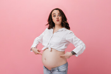 Blue-eyed brunette woman in white cropped shirt and jeans makes funny face. Pregnant lady points at her belly on pink background