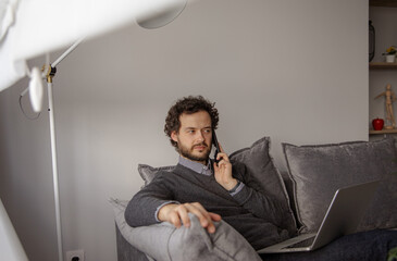 Fototapeta premium The guy solves serious questions over the phone. The man sits on a gray couch and talks on the phone. Work from home