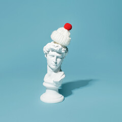 Male plaster bust with wool hat on the top of the head, minimal creative winter holidays inspired layout against pastel blue background. 