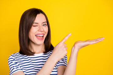 Photo of young attractive smiling cute woman blinking finger directing holding palm empty space advert isolated on yellow color background
