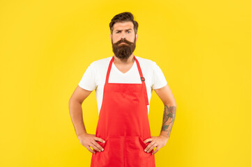 Confident serious man with arms akimbo in red apron yellow background, household