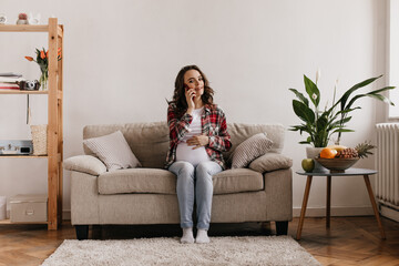 Cool pregnant woman in plaid shirt, white tee and jeans touches belly. Happy brunette girl talking on cellphone and poses on beige sofa