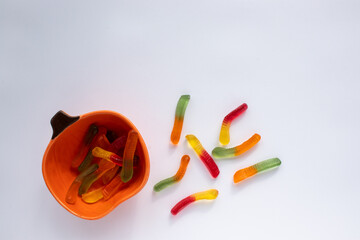 Halloween treats gummy worms in pumpkin bowl. Colorful jelly candies on white background. Top view,...