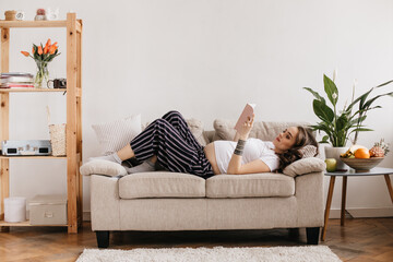 Curly calm pregnant woman lies on beige soft sofa in living room. Brunette girl reads pink book and rests. Portrait of young mother