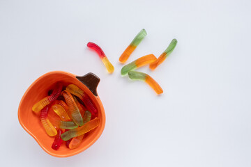 Halloween treats gummy worms in pumpkin bowl. Colorful jelly candies on white background. Top view,...