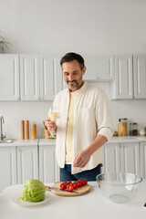 joyful man in white shirt holding glass of wine near fresh lettuce and ripe cherry tomatoes.