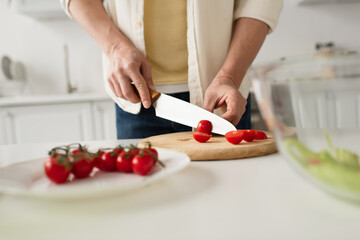 partial view of man cutting cherry tomatoes on chopping board on blurred foreground.