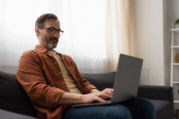 positive bearded man in eyeglasses sitting on couch and typing on laptop.