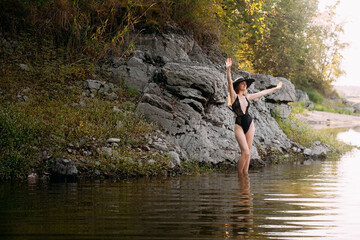 A young girl with long blond hair and a beautiful figure, in black openwork lingerie and a black wide hat, stands against the backdrop of a rock, on the banks of a picturesque river.