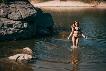 Fototapeta premium A young girl with long blond hair, a slender figure, in a black swimsuit jumps in the river against the backdrop of rocks and laughs.