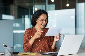 Businesswoman received happy news letter from bank, Hispanic woman with curly hair and glasses works inside modern office building, uses laptop.