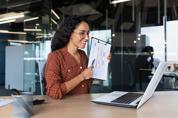 Business woman explains report indicators and financial results on video call, Hispanic woman smiles and looks at laptop web camera, female worker works inside modern office building with reports.