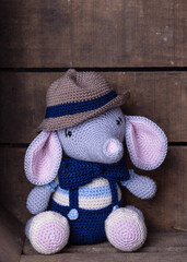 Cute gray and pink elephant with blue bow tie, blue pants and brown hat, in wooden box, wooden background, amigurumi toy, handmade, crochet, hobby, copy space
